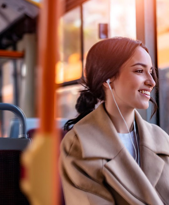 A young woman with dark hair smiles warmly, wearing earphones while seated on public transportation, sunlight streaming through the window.