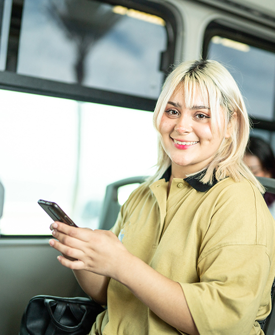 A smiling blonde woman in a tan shirt with black trim sits comfortably on a bus, holding a smartphone as light streams in.