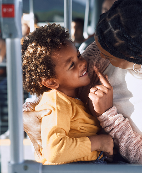 A joyful mother in a pink cardigan gently taps her smiling child's nose in a playful gesture while riding on public transit.