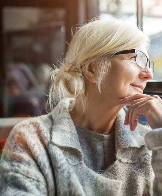 An older woman with white hair, in a cozy coat and glasses, sits by a window, gazing thoughtfully outside.