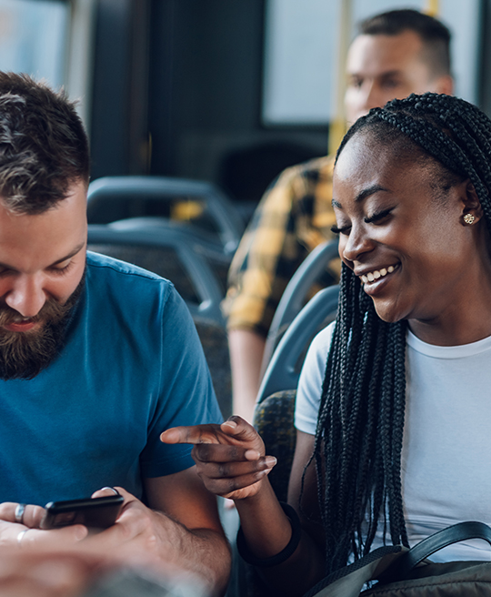 A young woman points towards a smiling man’s phone, both looking at the screen and laughing together while sharing a playful moment.