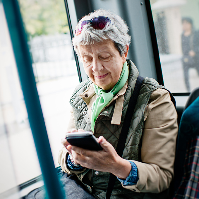 An older woman wearing a green scarf, beige jacket, quilted vest, and sunglasses on her head uses her smartphone on public transportation.